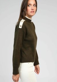 Pull en vert olive foncé avec un col montant, ourlet côtelé et accents blancs aux épaules agrémentés de boutons décoratifs. Texture tricotée.