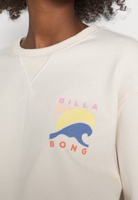 Sudadera color marfil con un gráfico que muestra una ola azul, un sol amarillo y acentos rosa. Hecha de un tejido suave y texturizado.