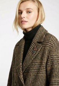 Manteau en laine à carreaux pied-de-poule marron et noir avec larges revers, agrémenté d'une broche décorative et d'un col roulé côtelé noir en dessous.