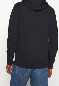 Sudadera negra con bolsillo tipo canguro, mangas y dobladillo acanalados. Textura suave, con una gran capucha y un diseño de espalda sin costuras, combinada con jeans azules.
