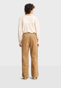 Blusa color crema con retro arricciato, abbinata a pantaloni in corda a coste color sabbia, a vita alta. Modello visto da dietro.