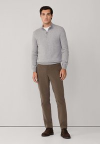 Hombre de pie con un suéter de cuello zip gris claro, pantalones marrones y zapatos de cordones marrones oscurecidos, contra un fondo claro y sencillo.