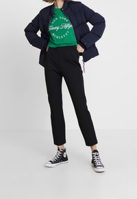 Mujer vestida con una chaqueta acolchada negra, camiseta verde de Tommy Hilfiger, pantalones negros y zapatillas Converse negras de caña alta, de pie con las manos en las caderas.