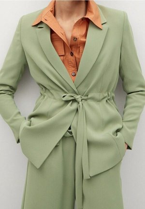 Jasnozielona marynarka wiązana w pasie i dopasowane spodnie noszone na otwartej pomarańczowej koszuli z rozpiętym kołnierzem, ręce w kieszeniach marynarki.