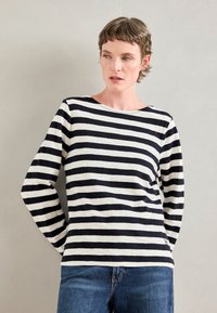 Kvinne med kort hår som har på seg en svart og hvitstripet langermet skjorte og blå jeans, står mot en ensfarget lys grå vegg.