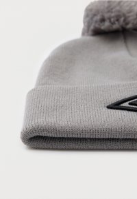 Salocīts gaiši pelēks adīts cepure ar pom-pom virsotnē un melnu izšuvumu piedurknē, uz balta virsmas.