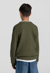 Maglione in maglia verde oliva con superficie testurizzata e polsini a costine, con scollatura rotonda e vestibilità comoda, abbinato a pantaloni corti in denim.