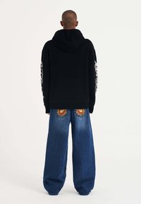 Felpa nera con orlo a coste e scritte bianche sulle maniche, abbinata a jeans blu larghi con ricami floreali sulle tasche posteriori.