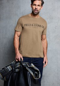 Světle hnědé bavlněné tričko s textem "FIELD & STONE CO." Model drží černou koženou tašku a kostkovanou košili v černé, šedé a žluté barvě.