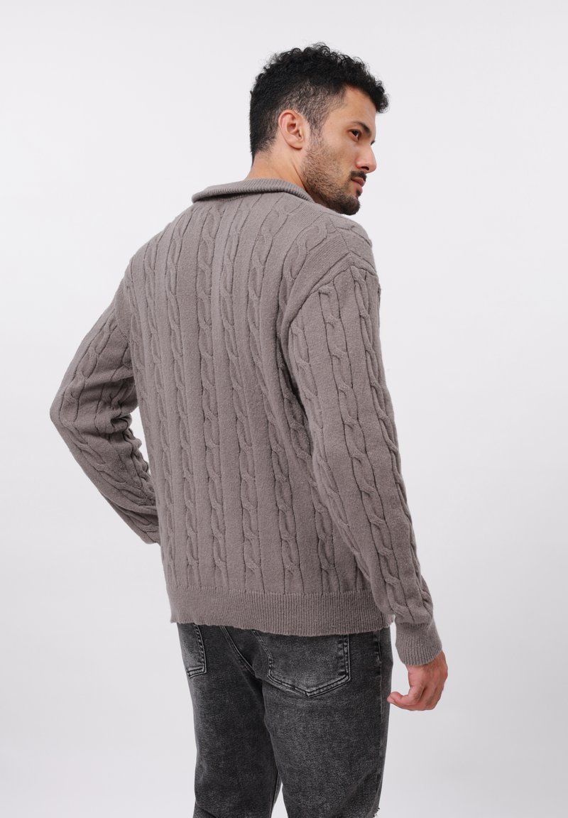 Mężczyzna w szarym swetrze z warkoczykiem i czarnych, wypłowiałych dżinsach, spoglądający przez ramię na tle białym.