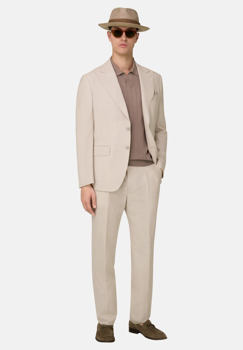 Homme en costume beige avec pantalon assorti, polo en maille marron, mocassins marron, chapeau fedora et lunettes de soleil rondes, debout avec une main dans la poche.