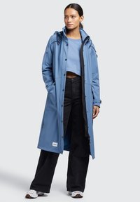 Abrigo largo azul impermeable con capucha, cierre a presión en el frente y ribetes negros. Lleva puesto un suéter azul claro y pantalones negros. Zapatillas blancas visibles.