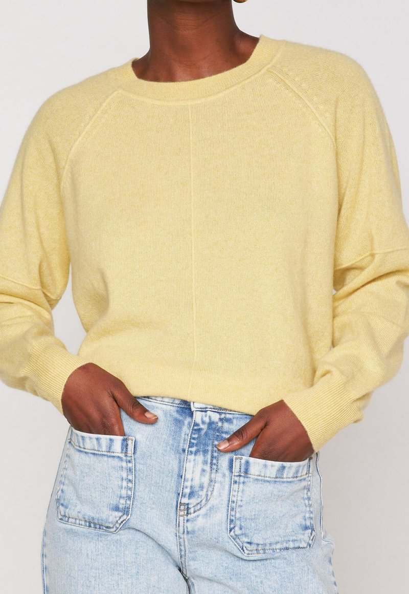 Personne portant un pull à manches longues jaune pâle avec des poignets côtelés et un jean bleu clair taille haute avec des poches plaquées à l'avant.