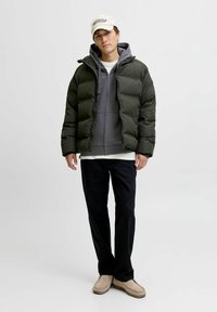 Chaqueta acolchada en verde oscuro con cierre frontal, llevada sobre una sudadera gris y una camisa blanca, combinada con pantalones negros y zapatos slip-on beige.