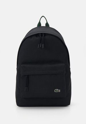 Svart stoffryggsekk med rund form, frontlomme med glidelås, justerbare polstrede stropper og en grønn Lacoste-logo på forsiden.