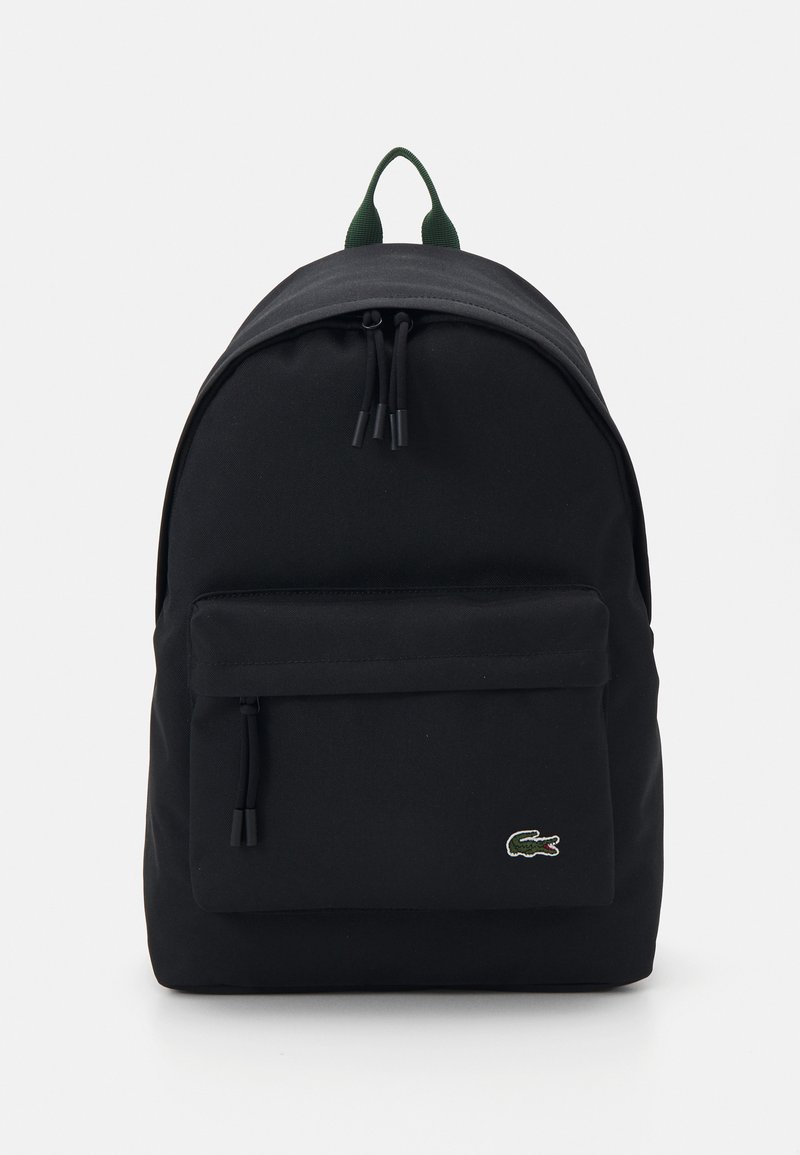 Čierny látkový batoh so zaoblenej forme, prednou vreckom na zips, nastaviteľnými polstrovanými popruhmi a zeleným logom Lacoste na prednej strane.