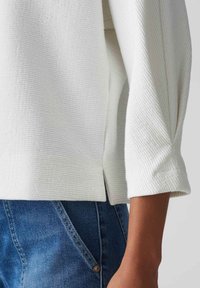 Haut blanc texturé à manches trois-quarts, avec une fente latérale. Associé à un denim bleu, mettant en valeur le mélange des matériaux et des styles.