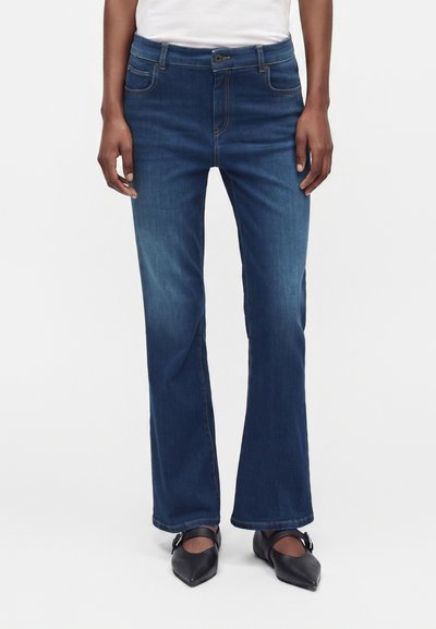 Personne portant un jean bootcut bleu foncé, un haut blanc et des chaussures noires à bouts pointus avec des brides à la cheville, debout devant un fond uni.