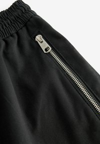 Gros plan sur un tissu noir avec une ceinture élastique et une fermeture éclair en métal argenté sur une poche.