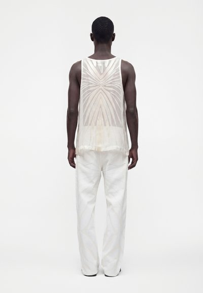 Homme de dos portant un haut sans manches blanc transparent avec des motifs géométriques et un pantalon blanc ample sur un fond blanc uni.