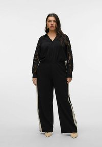 Completo nero che presenta una camicia bottonata con maniche lunghe in pizzo e pantaloni larghi con strisce laterali color crema. Indossato con scarpe beige a punta.