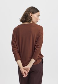 Maglietta marrone a maniche lunghe con un fit morbido, scollatura rotonda e texture morbida. Il retro presenta un leggero scollo a goccia ed è abbinata a pantaloni scuri.