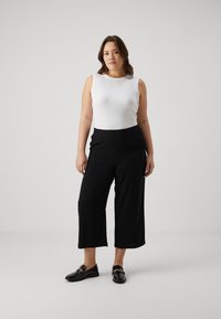 Débardeur blanc sans manches, pantalon noir à jambes larges, et mocassins noirs avec détail en chaîne. Tissu lisse, coupe décontractée, couleurs neutres.