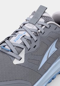 Grau sportlicher Schuh mit strukturiertem Obermaterial, hellblauen Akzenten, flachen Schnürsenkeln und verstärkten Ösen. Für Performance mit Dämpfung konzipiert.
