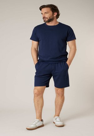Homme barbu portant un t-shirt bleu marine et un short, debout les mains dans les poches, regardant sur le côté sur un fond beige uni.