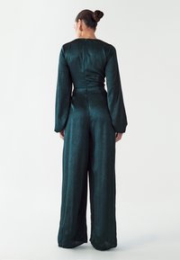 Grüner Satin-Overall mit langen Ärmeln, tailliertem Oberteil und weit geschnittenen Hosenbeinen. Rückseitiger Reißverschluss und strukturierte Stoffdetails.