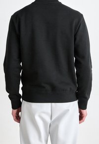 Osoba v jednoduchém černém dlouhém svetru a světlých šedých kalhotách, viděná zezadu na neutrálním pozadí.