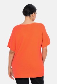 Světlé oranžové oversize pletené tričko s krátkými rukávy, které má žebrovanou strukturu a uvolněný střih, stylizované s černými kalhotami.