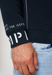 Sudadera azul marino con puños acanalados que presenta texto en blanco. El material es suave con una textura lisa y costuras claras contrastantes.