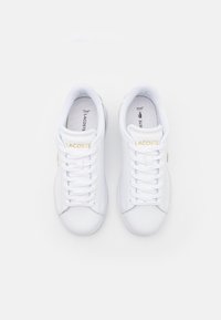 Adidași Lacoste albi cu un exterior din piele netedă, șireturi în față, branding auriu "LACOSTE" pe limbă și talpă din cauciuc texturat.