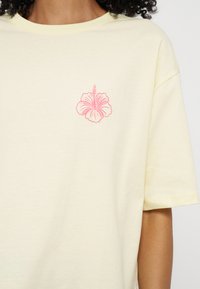 Personne portant un T-shirt jaune pâle avec un petit motif de fleur d’hibiscus rose sur la poitrine gauche.