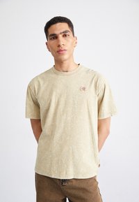 Camiseta de manga corta de color beige claro con un acabado texturizado y un pequeño parche de logo en el lado izquierdo del pecho. Corte relajado.