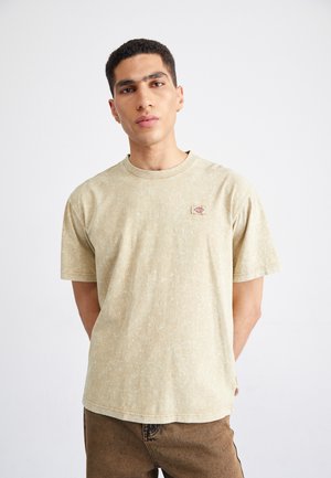 T-shirt corto a maniche corte color beige chiaro con una finitura testurizzata e una piccola etichetta con logo sul lato sinistro del petto. Vestibilità comoda.