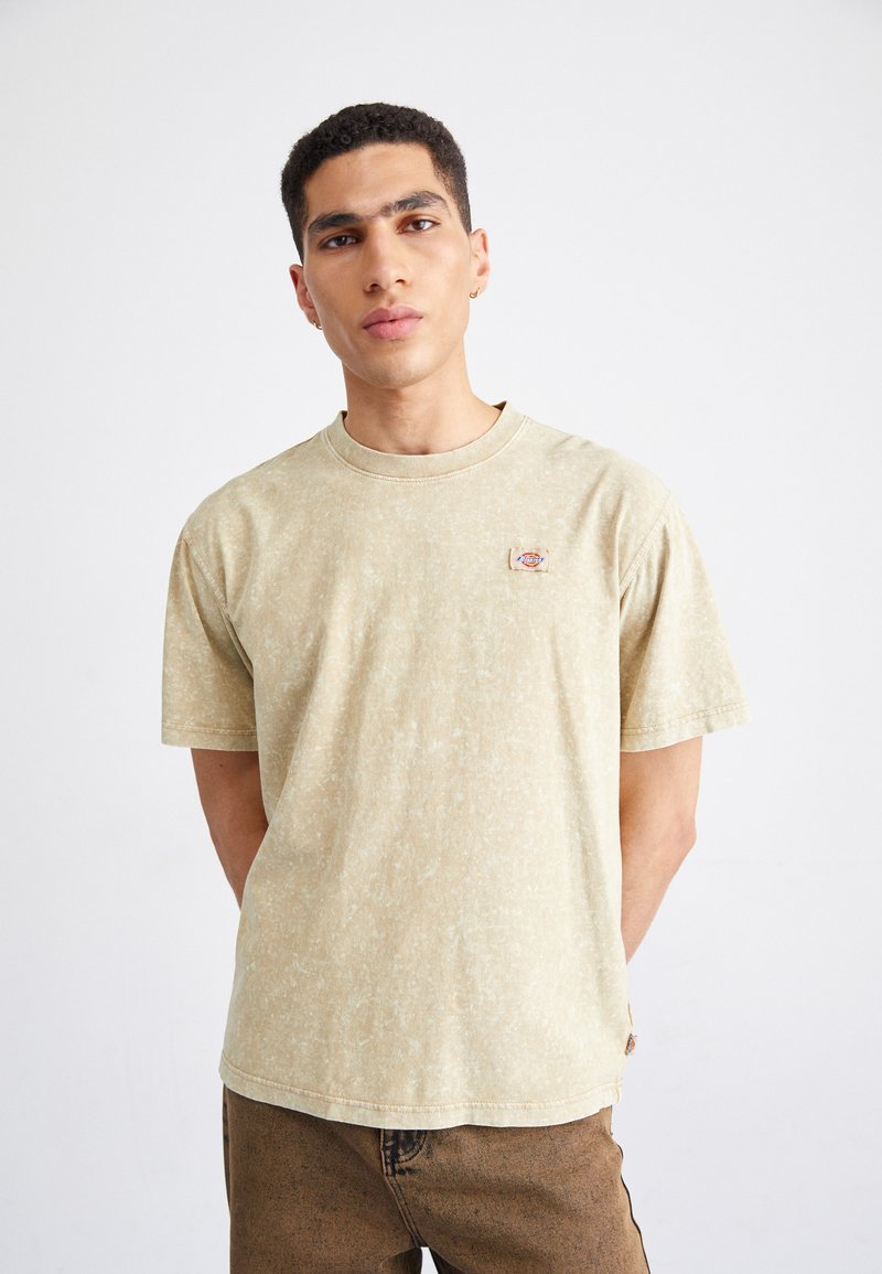 Camiseta de manga corta de color beige claro con un acabado texturizado y un pequeño parche de logo en el lado izquierdo del pecho. Corte relajado.