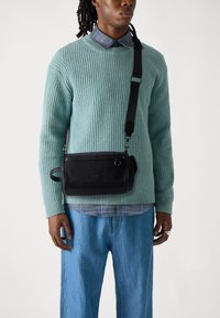 Borsa nera a spalla con una texture liscia, chiusura con zip e un piccolo manico, indossata sopra un maglione a coste blu chiaro e pantaloni di denim.