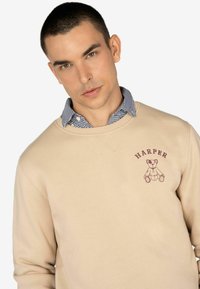 Sudadera beige de manga larga, con un gráfico de un oso marrón y el texto "HARPER" bordado en el pecho. Camisa de cuello debajo.