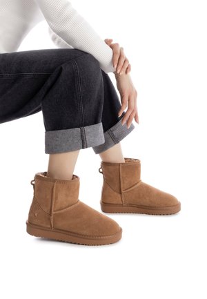 Botines de ante marrón con un suave forro interior, suela de goma plana y una lengüeta en la parte posterior. Llevados con unos jeans oscuros arremangados.