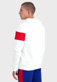 Haut à capuche blanc avec un accent rouge sur le bras gauche, doté d'une capuche avec cordon de serrage et de poignets côtelés, associé à un pantalon bleu. Texture lisse.