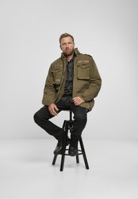 Olive green military-style jacket with multiple pockets, brown accents, and a textured interior. Worn over a black shirt and dark pants.