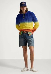 Άτομο που φοράει ένα μπλε, κίτρινο και πορτοκαλί gradient πλεκτό πουλόβερ, τζιν σορτς, λευκά αθλητικά παπούτσια, σκούρο μπλε καπέλο και γυαλιά ηλίου κρεμασμένα στο πουλόβερ.