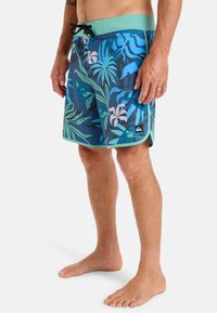 Herrarnas badbyxor med ett livligt tropiskt mönster i blått, grönt och rosa, med en enfärgad grön midjeband och svart logomärke.