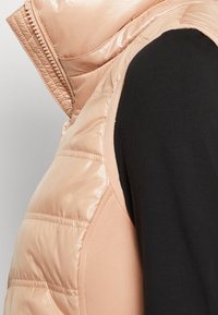 Jasno beżowa puchowa kamizelka z wysokim kołnierzem, z połyskliwym wykończeniem i poziomym pikowaniem. Akcentowana czarnym materiałem pod spodem.