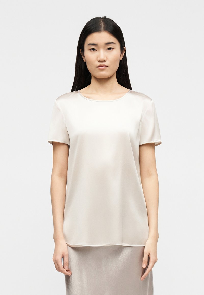 Γυναίκα με μακριά σκούρα μαλλιά που φοράει ένα κ οντομάνικο, ανοιχτό μπεζ σατέν μπλουζάκι και φούστα στον ίδιο τόνο, στέκεται μπροστά σε απλό ανοιχτό φόντο.