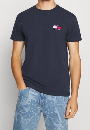 Mężczyzna ubrany w granatowy t-shirt Tommy Jeans i jasnoniebieskie jeansy w wzory, z jedną ręką w kieszeni, na jednolitym tle.