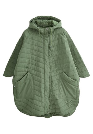Personne portant une veste matelassée verte avec de grandes poches, un jean bleu déchiré, des baskets blanches et une montre-bracelet en or, debout sur un sol en bois.
