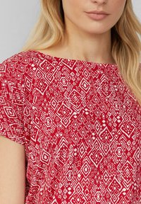 Rood stoffen top met een brede halslijn, voorzien van een geometrisch wit patroon van diamanten en vierkanten. Korte mouwen en een lichte textuur.